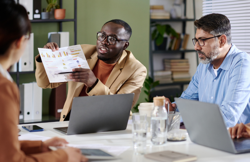 Colleague presents a printed chart with graphs to teammates during a meeting, discussing ROI at a table with laptops in a modern office.