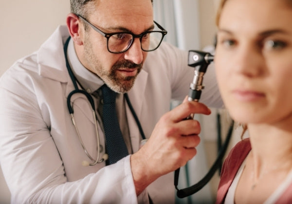 A doctor wearing a white coat and stethoscope examines a female patient’s ear with an otoscope during a consultation.