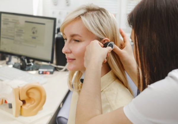 An audiologist performs an ear examination on a smiling female patient using an otoscope, with medical equipment and a computer displaying audiology software visible in the background. A model of a human ear is also seen on the desk.