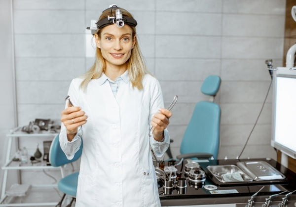 Female ENT surgeon in a white coat and headlamp, holding medical instruments, standing in a modern examination room with specialist equipment and a patient chair in the background.
